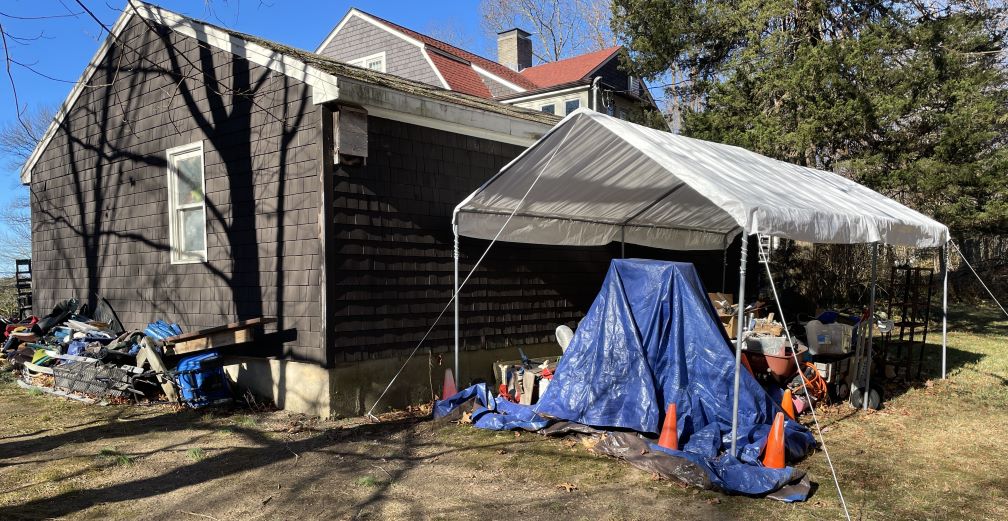 A brown shingled building. On one side is a large pile of stuff including some tarps, a contraption with three skis and a platform, a blue wheelbarrow on its side, and a wooden teeter totter. On the other visible side there is a white canopy with aluminum poles and white ropes going at an angle out to stakes in the lawn. Under the canopy is a structure underneath a blue tarp with some highway cones holding down the tarp, and behind that you can see a red wheelbarrow full of junk, some boxes with more junk, a hand truck, an empty shelving unit, and an orange tangle of hosing.