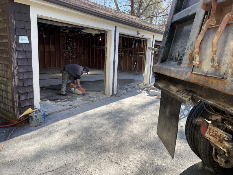 Angled shot of the interior of the garage; a bicycle is hanging on the rear wall. A workman is holding a cement saw cutting a slice through the garage floor. The rear of a large dump truck is visible to the right.