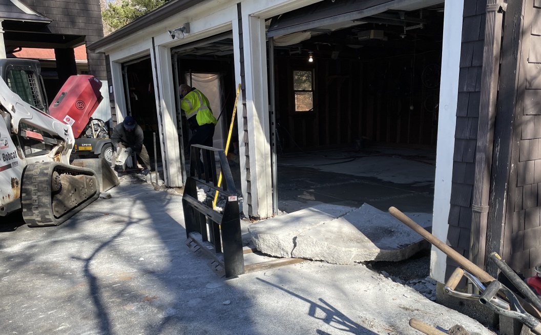 A Bobcat tractor with a front loader attachment on. A worker is placing a concrete block into the front loader. To the right there is a forklift attachment (not attached), with some much larger (4 ft. square) concrete blocks half in/half out of the garage next to it.