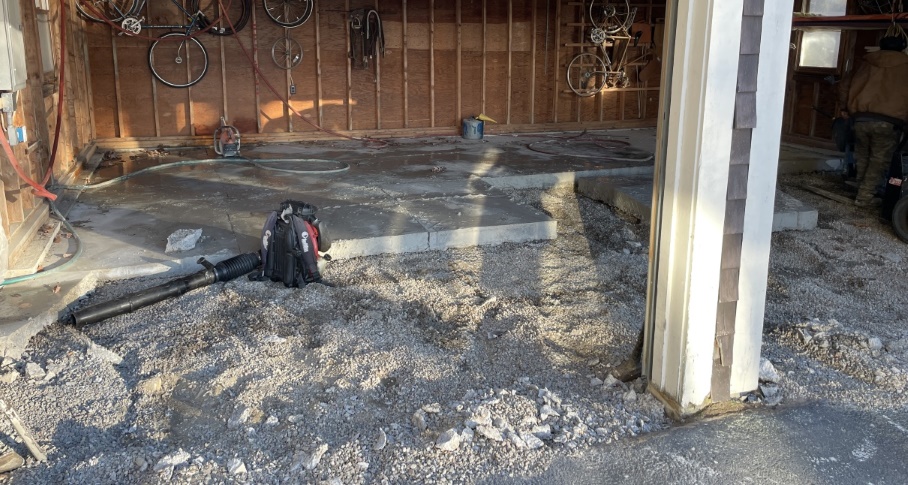 A slightly angled shot of one bay of the garage. You can see two bicycles and some bicycle wheels hanging on the rear wall. About the front half of the concrete blocks have been removed, revealing gravel beneath. The rear half has had slices cut through the concrete. There are some machines and garden hoses; the floor is wet.