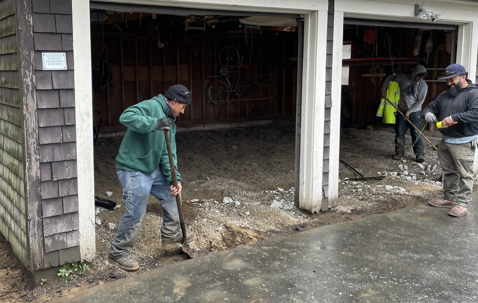 Angled shot of the garage with two workers using shovels and one doing something with yellow string for 'leveling' (it wasn't level: they gave it a slight slant, but they needed to make the slant the same everywhere.)