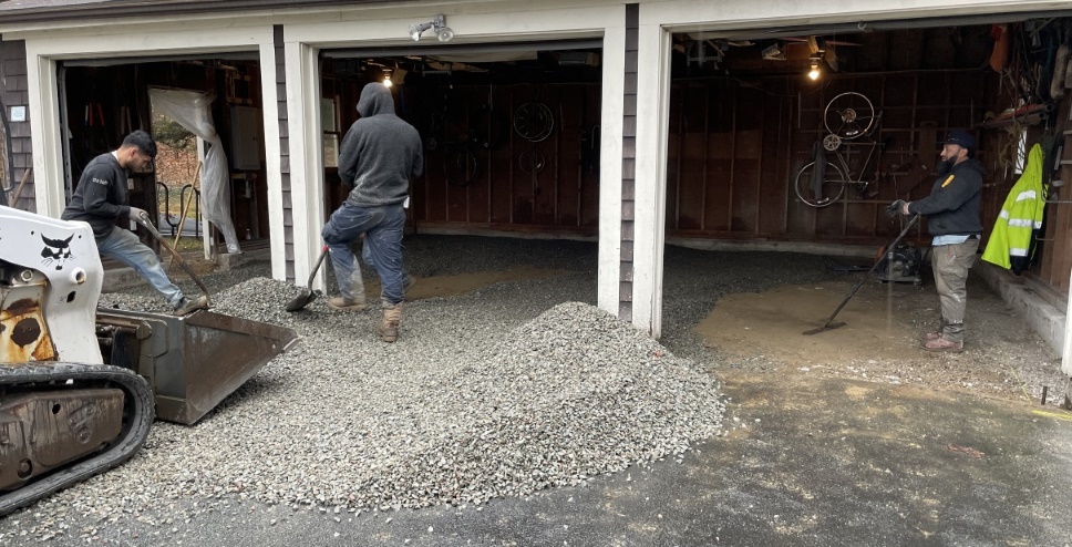 Slightly angled shot of the garage with a partial Bobcat machine on the left. There is a bunch of gravel in the rear of the garage and also in a big pile on the driveway, and also some dirt patches yet to be filled in. Two workers are wielding shovels and one is standing and watching.