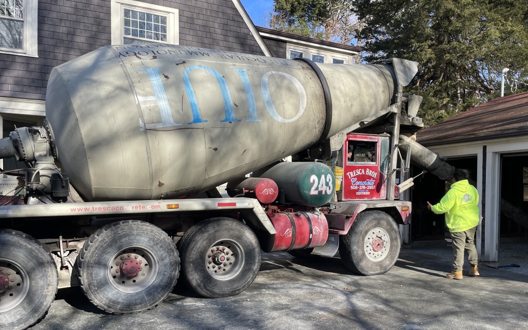 A cement truck from Tresca brothers, with a red door on the cab, and 'OUIF' printed (upside down from this vantage) on the large cone shaped cement mixing vessel. There are 4 tires visible on the side, holding up the heavy cement on the truck, and a few smaller cylindrical vessels, one of which is green and has the numbers 243 blazoned on the end. A worker in a bright yellow jacket stands by the door and speaks to the driver.