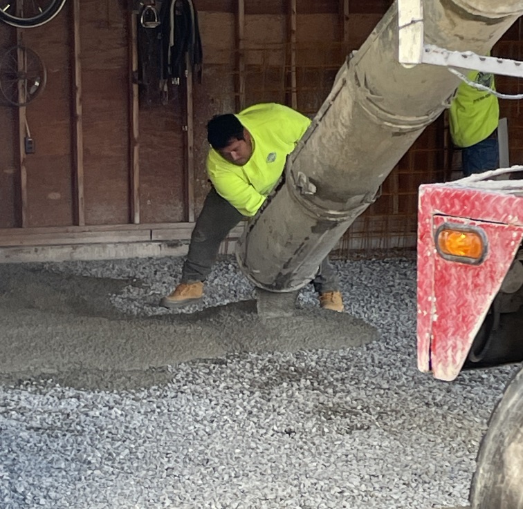 A worker in a bright yellow shirt, standing on the gravel inside the garage, guiding the cement pouring out of the tube at the front of the cement truck. A bit of cement truck is visible in the corner of the photo.