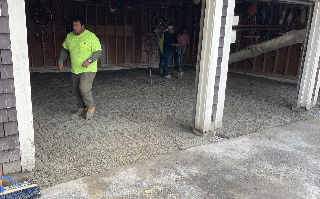 A worker in a bright yellow shirt and gray pants walks on freshly poured cement with rust colored six-inch square mesh reinforcing steel bars. In the background are some other workers with shovels and the cement extruding tube.