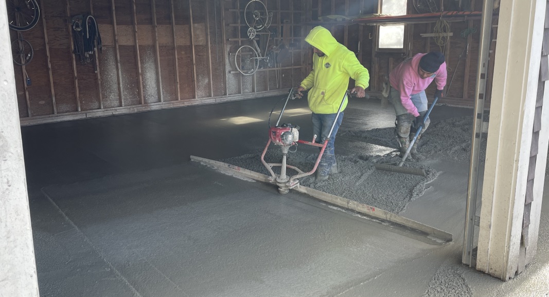 An angled shot of the garage. The rear half has smooth-looking cement. In the front half a worker in a bright yellow sweatshirt is pulling a contraption across the cement while a worker in a pink sweatshirt spreads the cement behind him. The contraption has a 10 foot wide blade and a motor mounted in the center with handles.