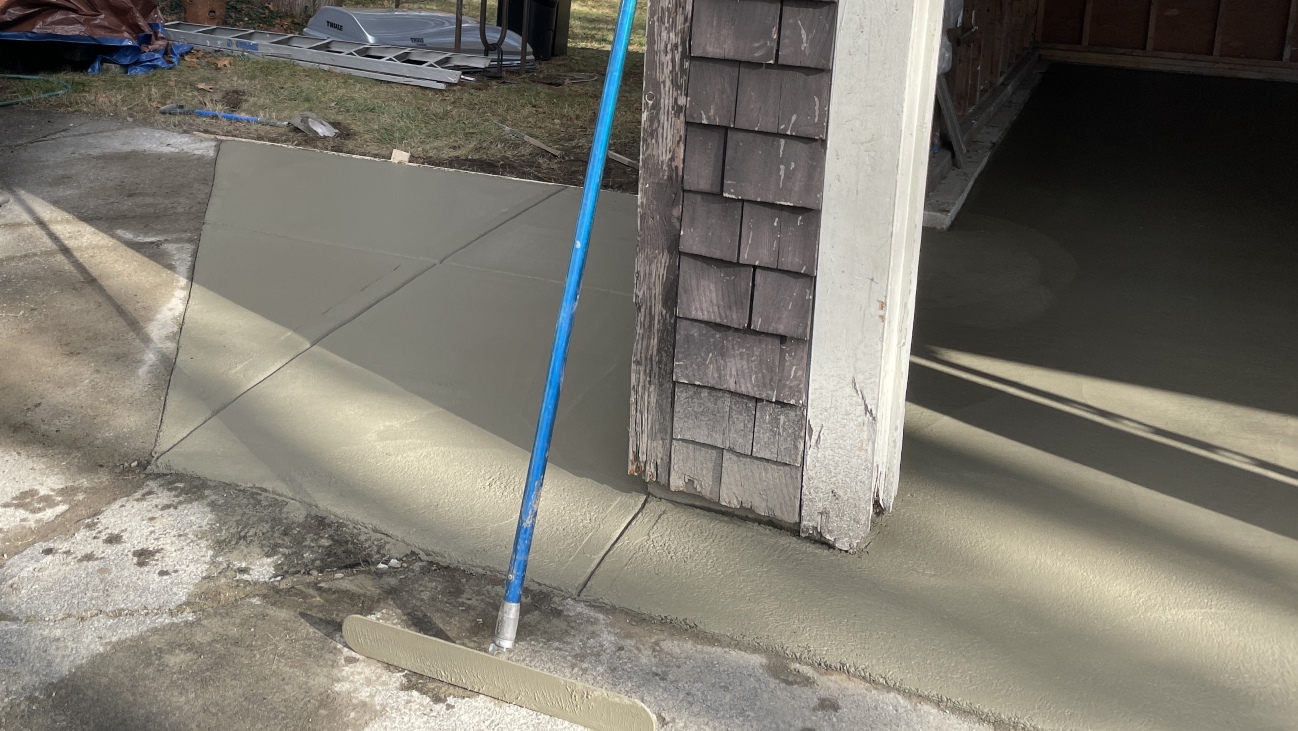 The far left hand side of the garage, interior seen as flat wet cement from before. To the left of the garage, outdoors, there is a trapezoidal piece of freshly poured and smoothed cement that abuts the asphalt driveway and an older piece of sidewalk at an angle. Behind the new cement is a grassy patch with much junk on it including a ladder. In front of the cement, leaning against the garage wall is a blue handled cement smoothing tool.