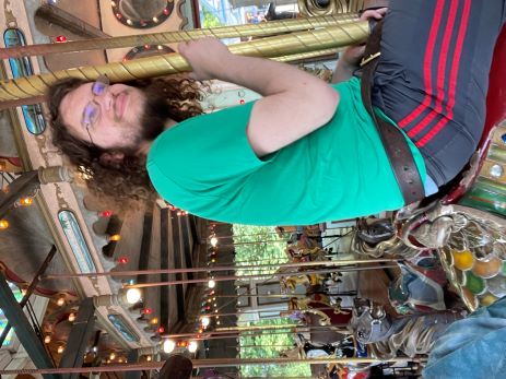 A sleepy looking young man in a green shirt and black shorts with red vertical stripes along the outside seam, sitting on a merry-go-round horse, leaning his head against the gold-colored pole in front of the saddle. There are many other merry-go-round horses visible in the background. His long brown hair and short scraggly beard are unkempt.
