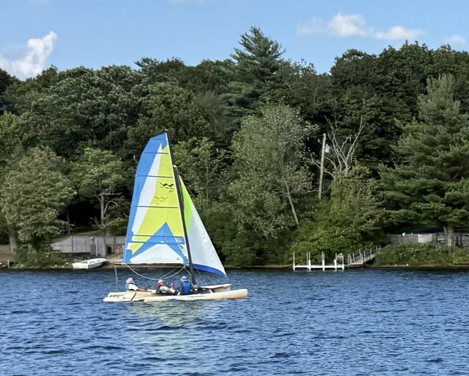 A sailboat, with a yellow hull and white, blue, and yellow sails is in a slightly wavy lake passing a dock with a variety of trees in full leaf behind that. The sky is blue with little wispy clouds. Three people can be seen on the boat. You can sort of tell it's a multihull, if you squint.