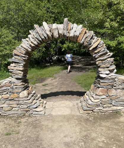 A nearly circular arch made of flat stones of a few inches wide in front of a tree-lined path. A person is walking down the path 100 or so feet away.
