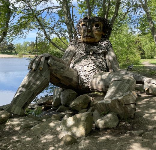 A large wooden sculpture of humanoid creature next to a pond, lounging against some trees with some rocks at its feet.