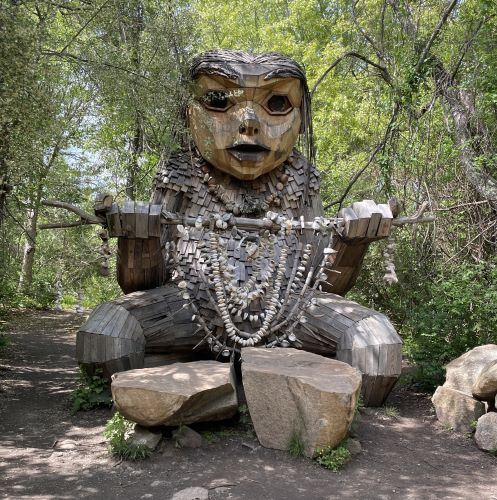 Front view of a large wooden sculpture of a humanoid creature kneeling in front of two flat top rocks. It looks female, with shingles covering its torso while its legs and arms have boards which are more smoothly connected. It is holding a stick between two outstretched arms. There are loops of shells and rocks descending from the stick.