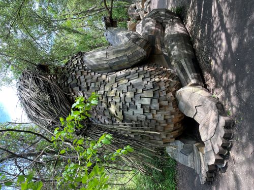 Rear view of a large wooden sculpture of a kneeling humanoid creature. It has long hair constructed of some kind of reeds, which is tied back with some kind of vine. It's torso is covered with shingles. You can see the feet under its rear end, which have great detail including a gap where the arch of the foot should be and toenails.