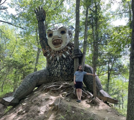 Front view of a large wooden sculpture of a troll on top of a small rise. Also included is a white man wearing a blue shirt and black shorts for scale. The troll is at least three times as tall as the man. The troll has a white painted face with its mouth wide open showing 4 large teeth. One is raised overhead and the other hand is holding a tree. Its legs are splayed out in a straddle. It looks like it's having fun sliding down the hill.