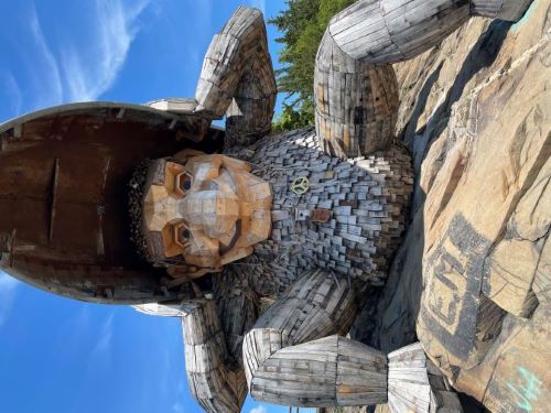 Front view of a large wooden sculpture of a humanoid shaped creature sitting on rocks. The arms and legs are made of
				  a set of boards smoothly connecting
				  to each other, while the torso is covered with shingles.  The face has a very large nose and chin. It is holding a red canoe-shaped boat above its head.