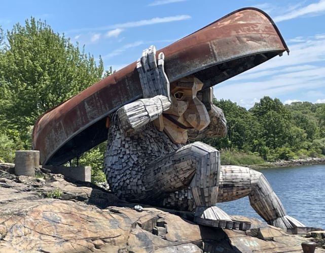 Side view of a large wooden
				  sculpture of a humanoid shaped
				  creature sitting on rocks by some
				  water. The arms and legs are made of
				  a set of boards smoothly connecting
				  to each other, while the torso is covered with shingles. The face has a very large nose and chin. It is holding a red canoe-shaped boat above its head.