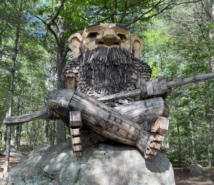 Front view of a humanoid wooden sculpture, seated crosslegged on a large rock. This one has little sticks evoking a beard and mustache below its very large nose. It has shingles for its torso and more smoothly connected small planks for its legs and feet. It is holding a pole.