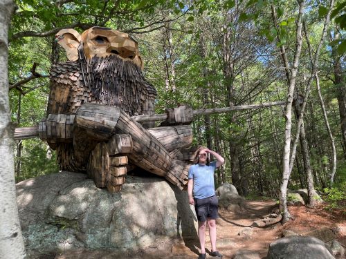 Angled view of a humanoid wooden sculpture, seated crosslegged on a large rock. This one has little sticks evoking a beard and mustache below its very large nose. It has shingles for its torso and more smoothly connected small planks for its legs and feet. It is holding a pole, at the far end of which is a small bird house. There is a young white man in a blue shirt and black shorts standing next to the rock for scale; the seated sculpture is easily twice his height.